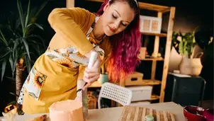 A woman with vibrant pink hair is icing a cake in a home kitchen, with macarons on a wooden board nearby